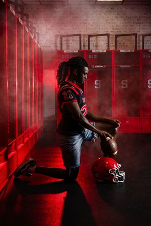 Football player in red uniform kneels in smoky locker room holding helmet and ball under dramatic red lighting.