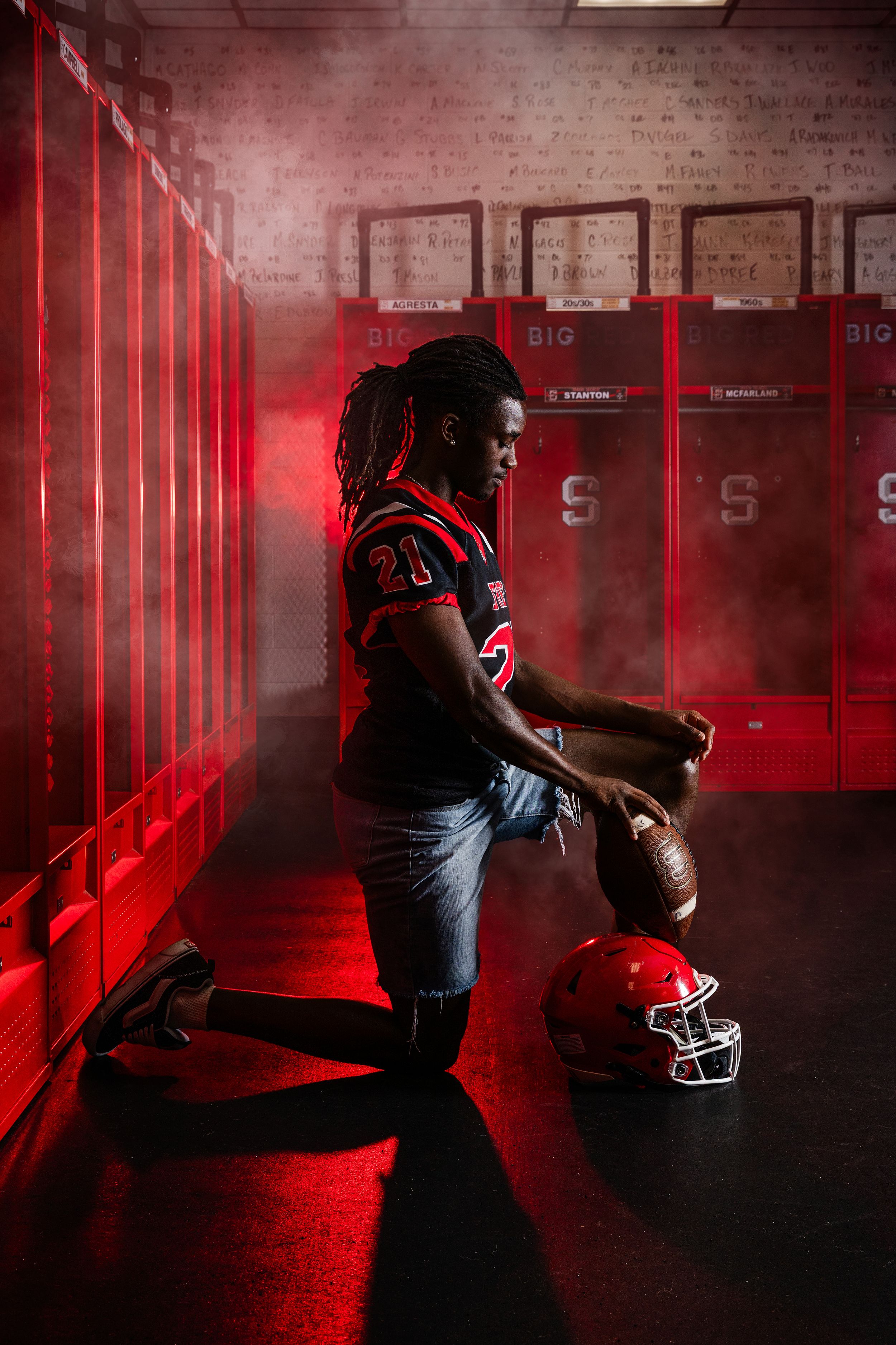 Football player in red uniform kneels in smoky locker room holding helmet and ball under dramatic red lighting.