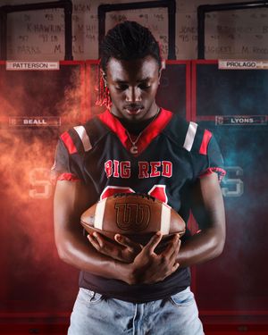 Football player in Big Red jersey holds Wilson football, dramatic red and blue smoke background.