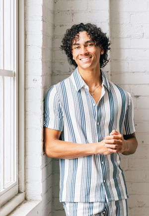 Smiling young man with curly hair wearing a blue striped short-sleeve button-up shirt, leaning against a white brick wall.