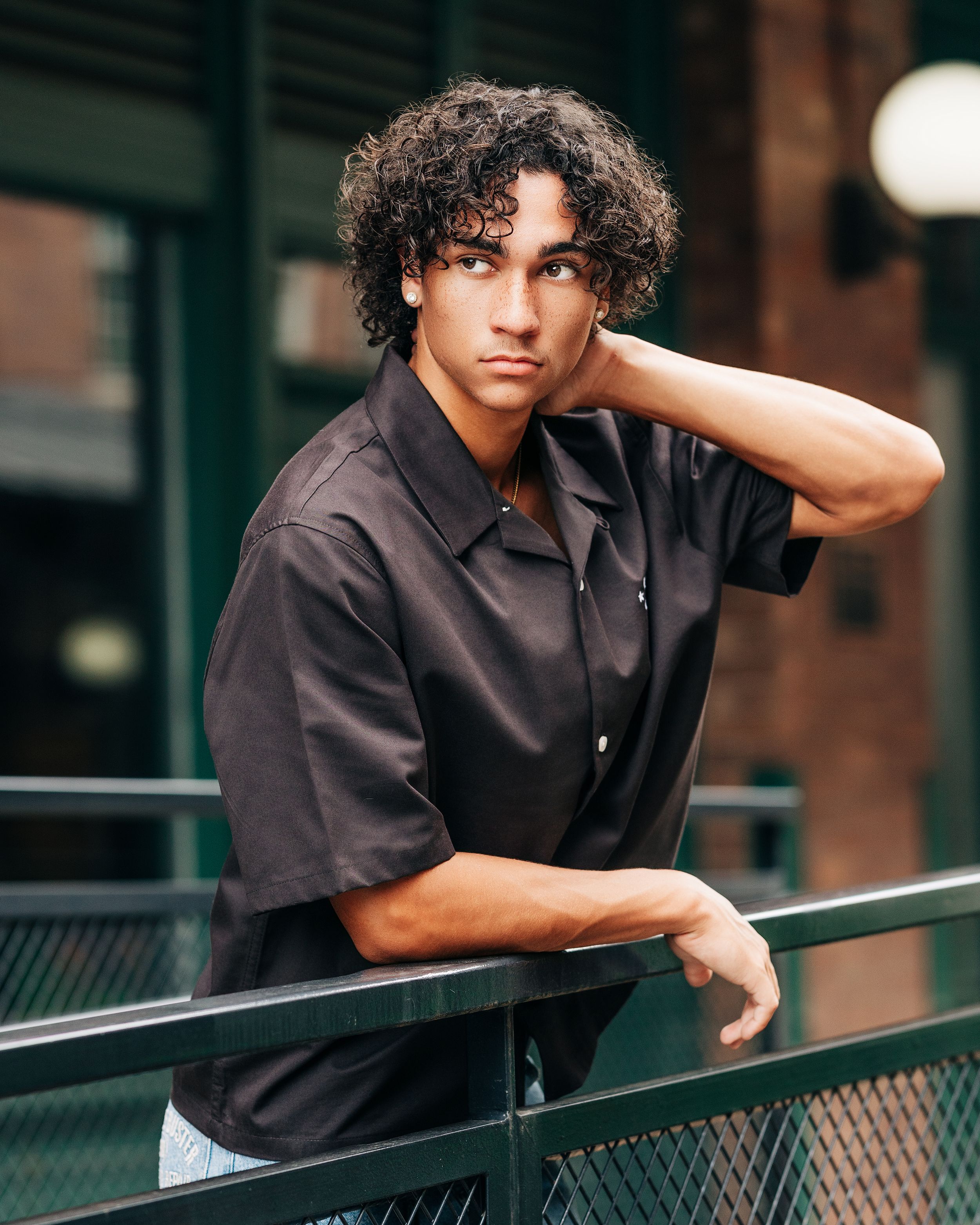 Young man with curly hair in black shirt leaning on railing in urban setting.