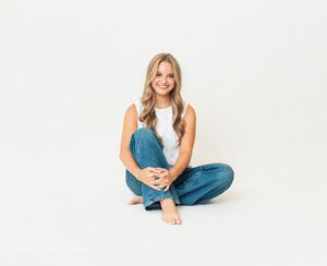 Young blonde woman sitting cross-legged on white background, wearing white top and blue jeans, smiling warmly.
