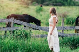 Young woman in white dress standing by wooden fence in green pasture with dark horses grazing nearby.