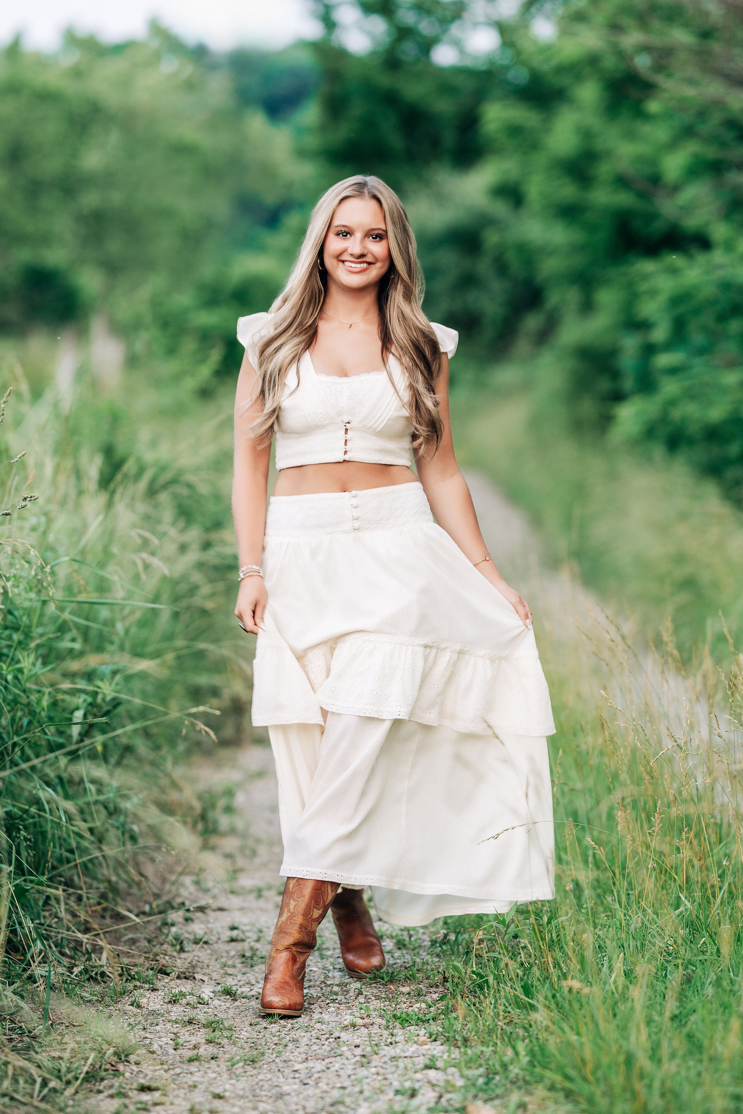 Young woman in white crop top and tiered maxi skirt with brown boots smiling on a scenic green nature path.