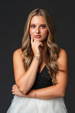 Young woman in black and white dress posing thoughtfully with hand on chin, wearing bracelets against dark background.