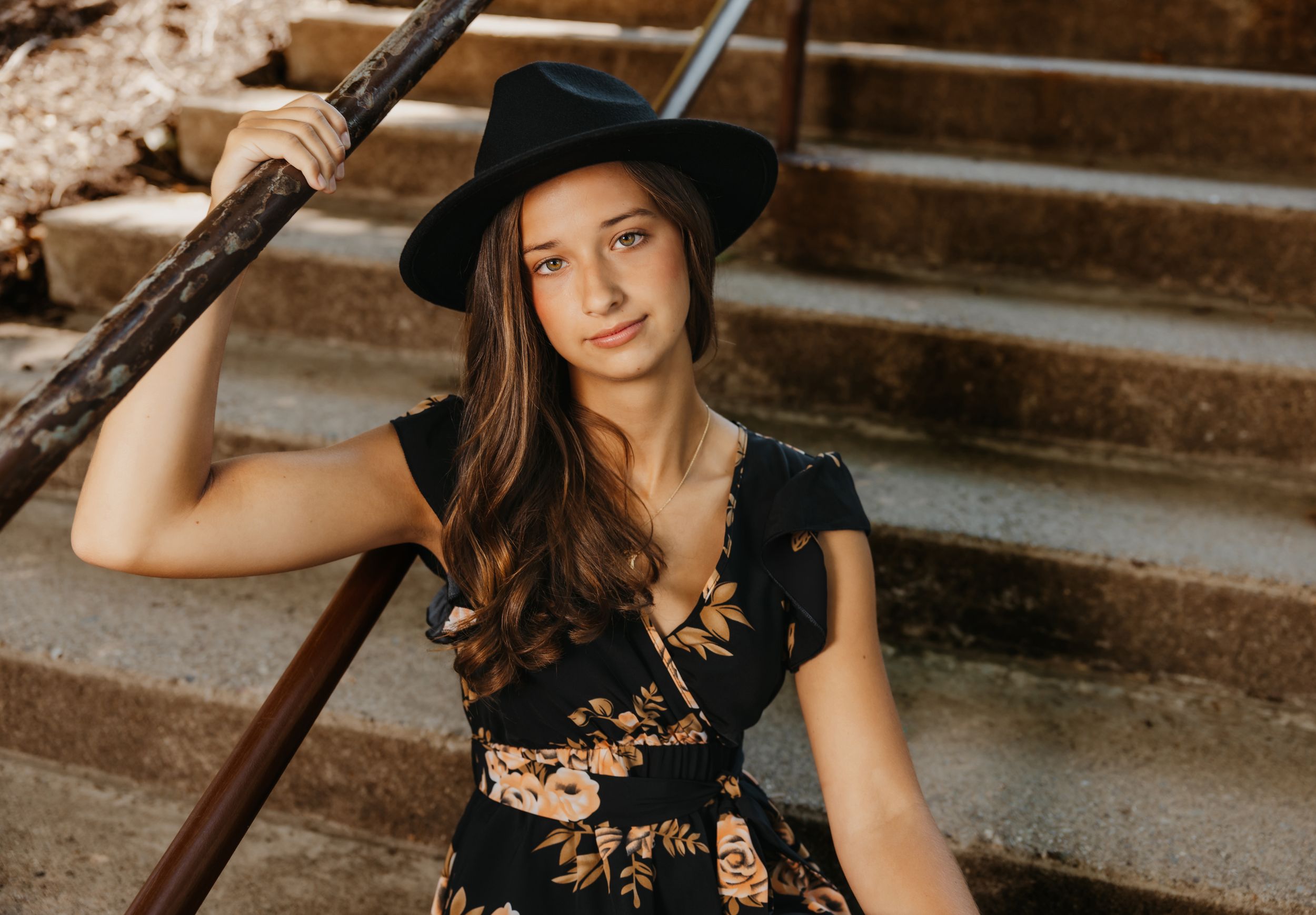 Portrait on stone steps featuring black hat and floral dress in warm sunlight.