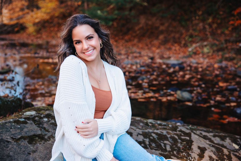 Portrait of someone in a white cardigan sitting on rocks near autumn leaves and water.
