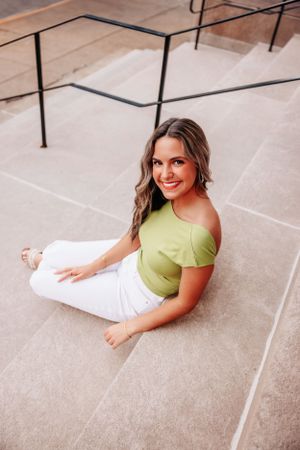 Woman in lime green crop top and white pants sits on concrete steps outdoors.