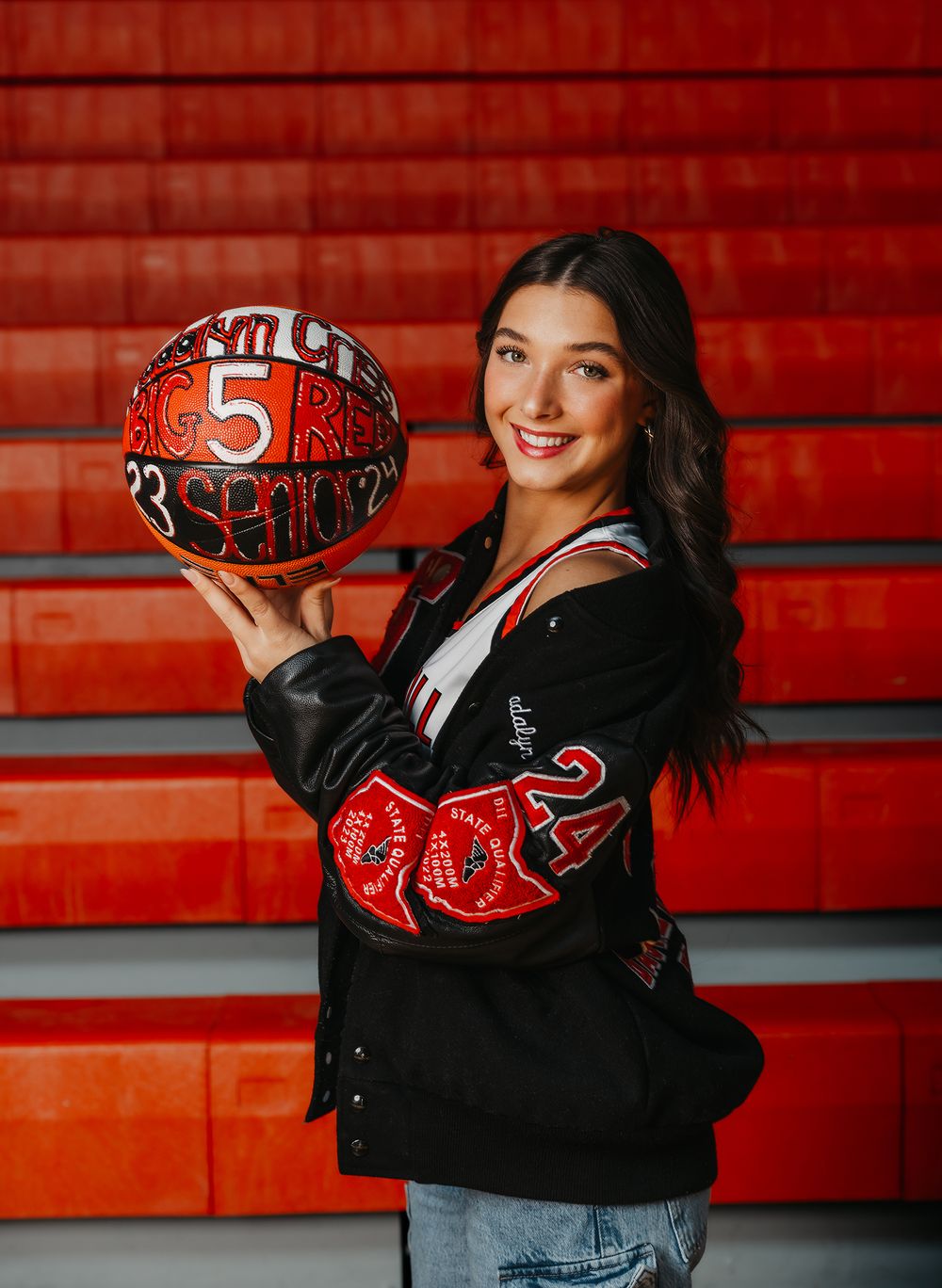 Basketball player holding red decorated ball while wearing varsity jacket against red bleachers.