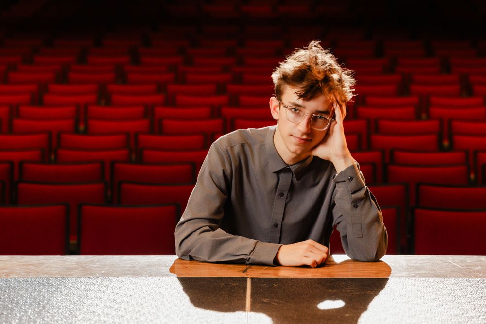 A person sits alone at a desk looking stressed in an empty theater with red seats in the background.