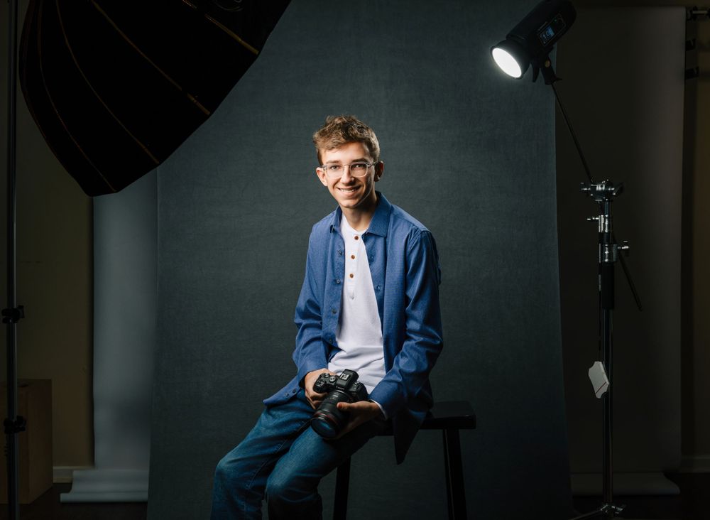 Photography studio setup with lighting equipment and backdrop showing a seated subject in denim and blue attire.