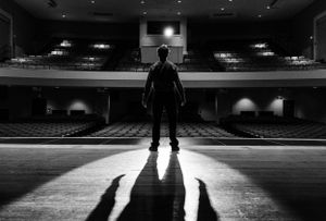 A silhouetted figure stands alone on an empty theater stage casting long shadows across the dramatic spotlit floor.