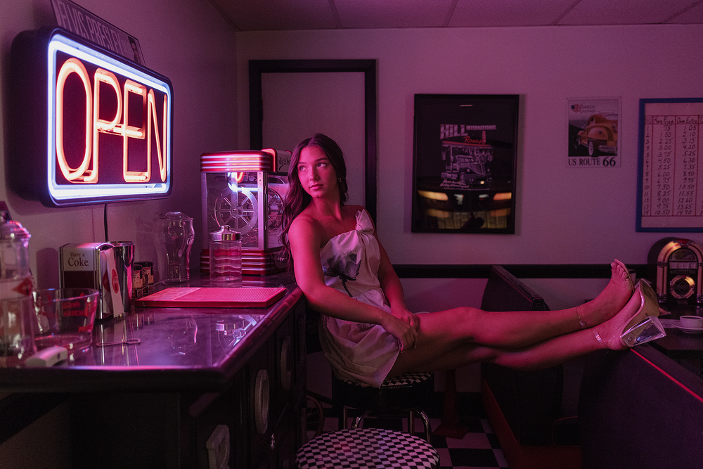 Person lounging on bar counter with neon open sign creating moody pink lighting atmosphere.