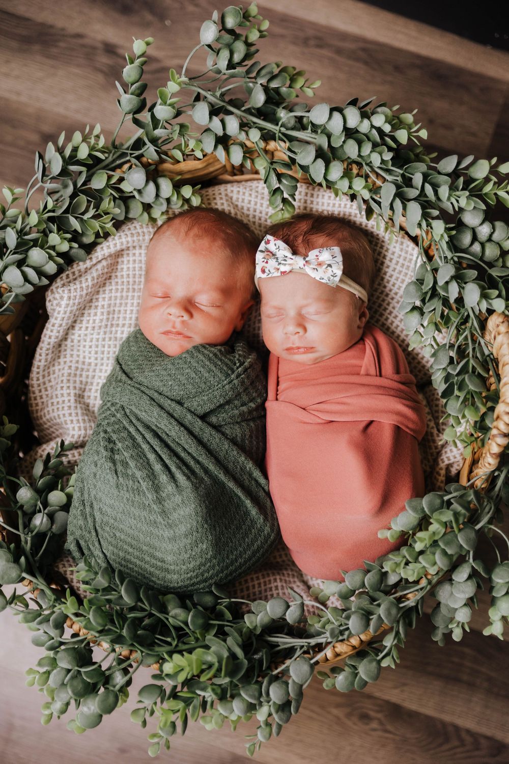 Newborn twins swaddled in green and coral wraps lay side by side surrounded by eucalyptus garland in a rustic setting.