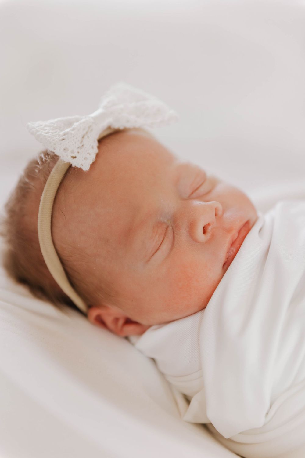 A sleeping newborn baby wrapped in white swaddle blanket with a delicate headband.