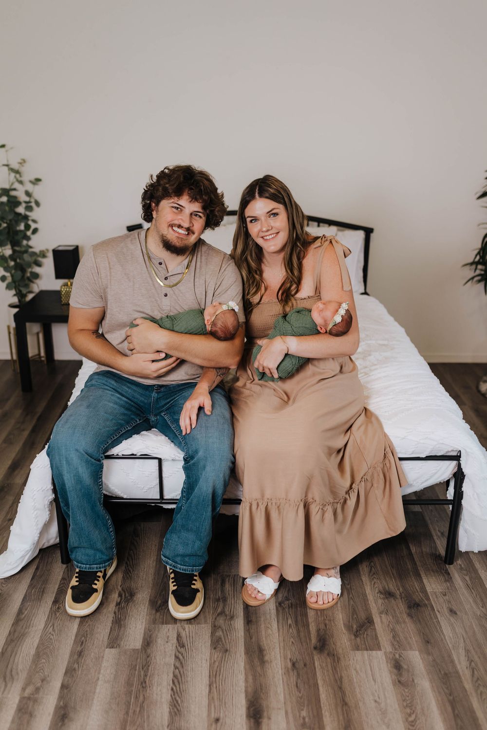 A couple sits together on a wooden floor against a white bed in a minimalist bedroom setting.