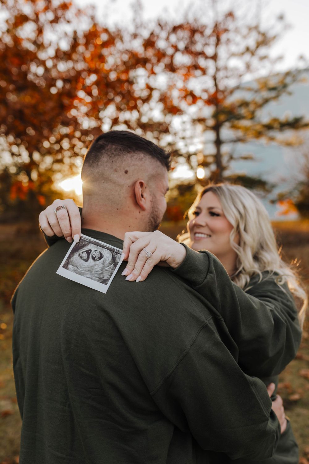 A couple embracing outdoors in autumn while sharing ultrasound photos.