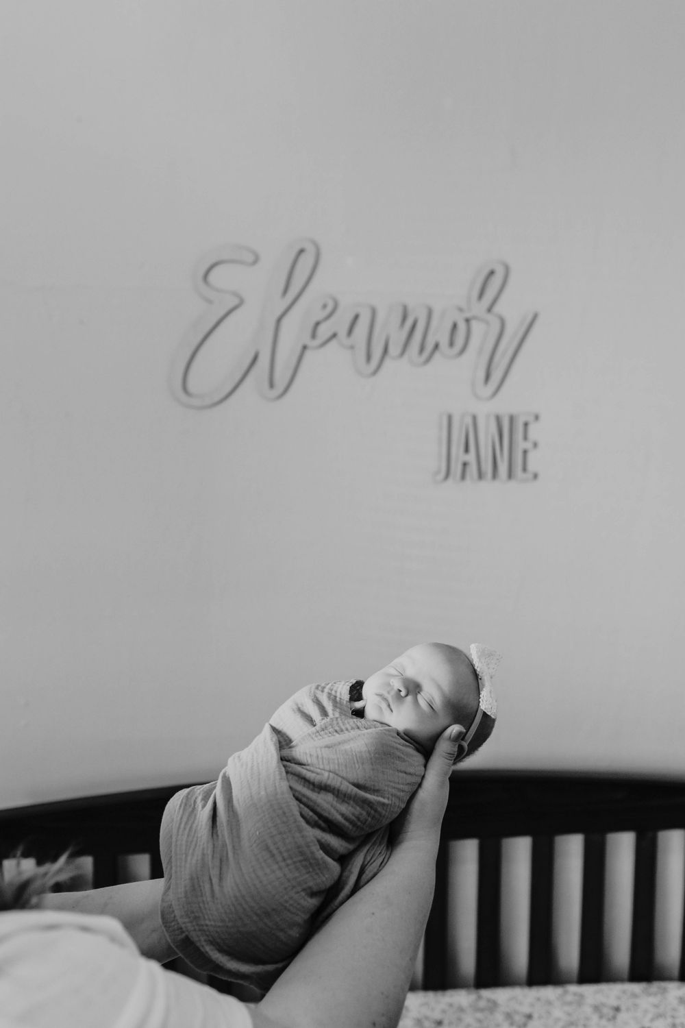 A newborn baby wrapped in a gray swaddle blanket lies in a wooden crib beneath handwritten text on the wall.