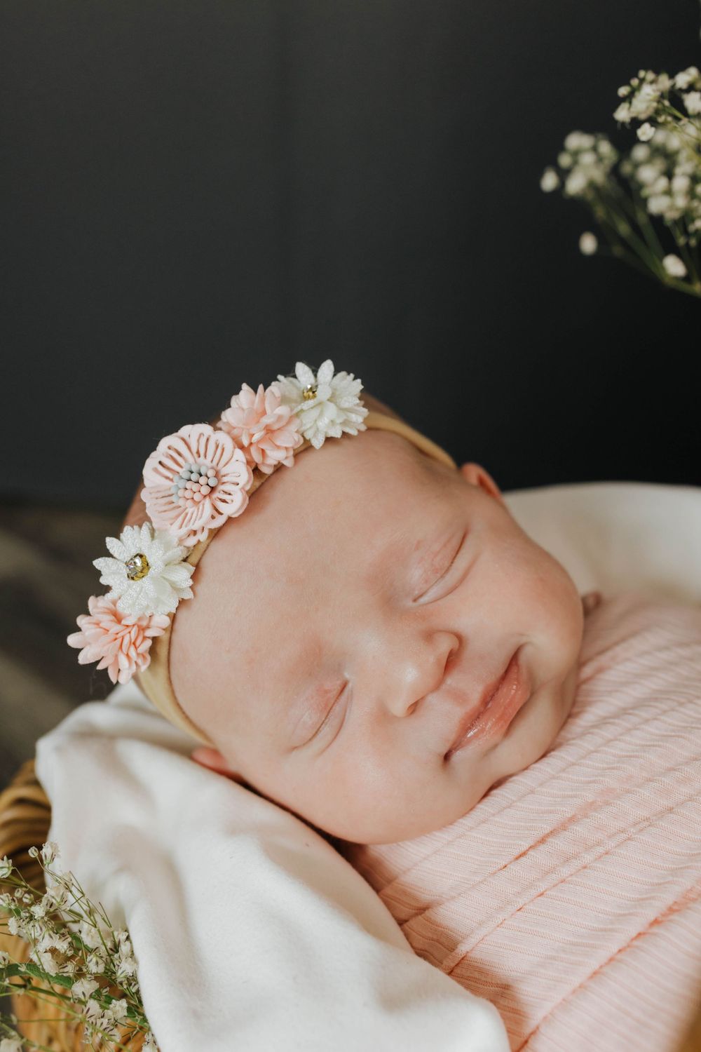 Sleeping newborn wearing delicate floral crown headband with pink and white flowers rests on soft blanket.