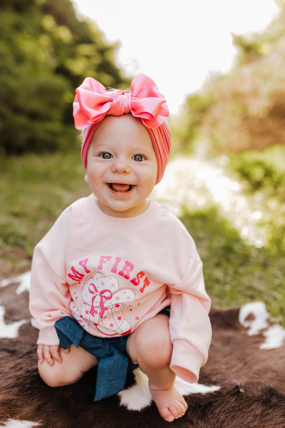 Baby wearing pink bow headband and white shirt sits on brown blanket outdoors in natural lighting.