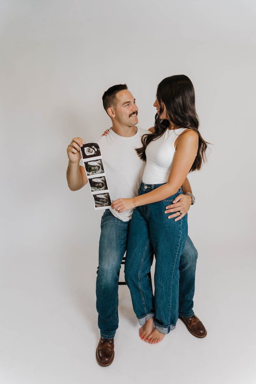 A couple in jeans shares a tender moment while holding an ultrasound photo against a white background in a pregnancy announcement.