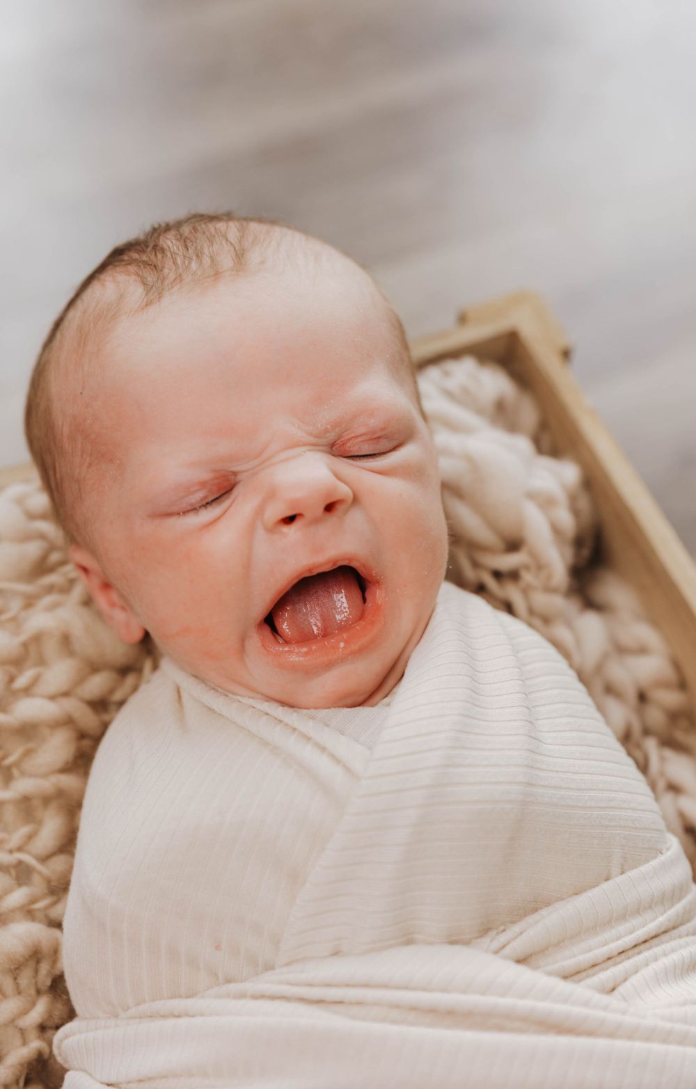 Newborn baby yawning while wrapped in white swaddle and lying on soft beige blanket in wooden crate.