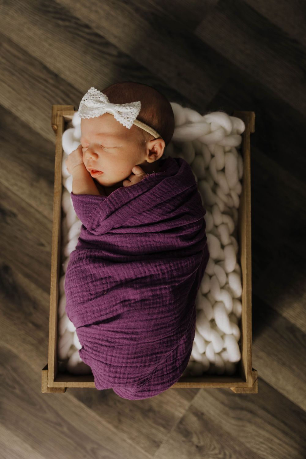 Sleeping newborn swaddled in purple muslin wrap with white bow headband resting in wooden box.