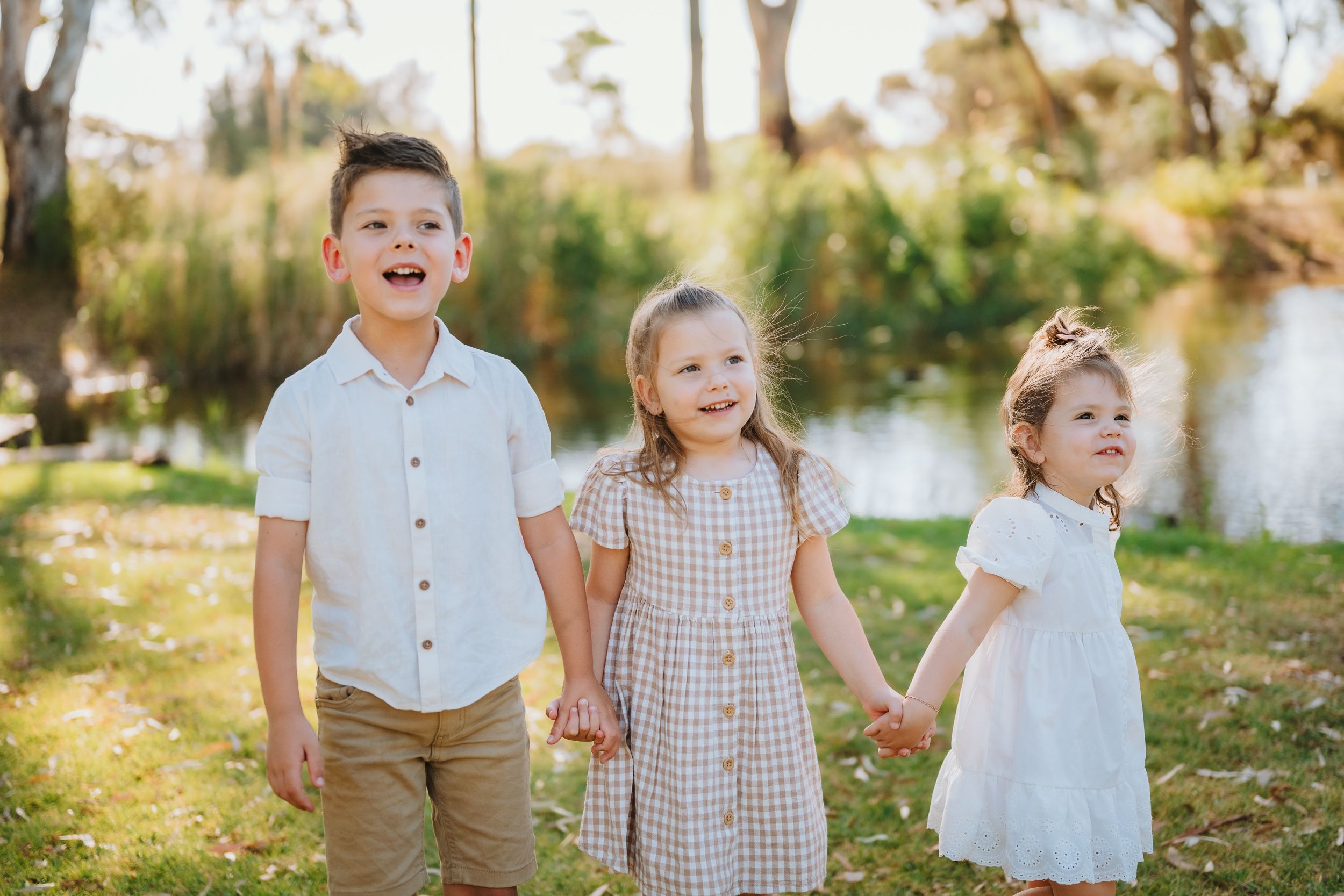Three children hold hands while standing together outdoors in front of a pond on a sunny afternoon.