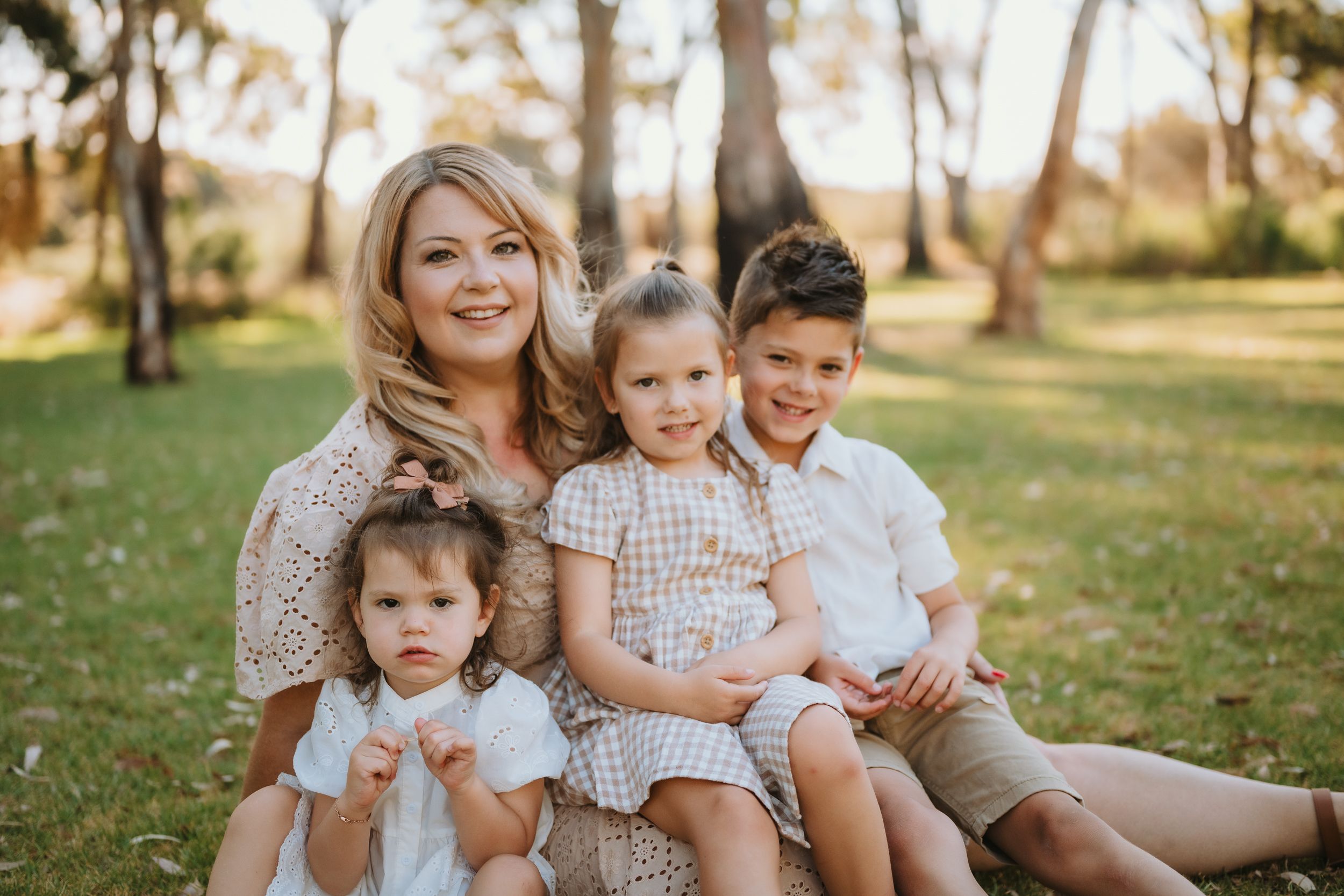 A family group sits together outdoors on grass in a sun-dappled wooded setting wearing coordinated neutral-colored outfits.