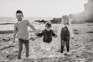 Three children hold hands and run joyfully together on a beach in black and white.