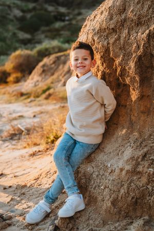 A young child in a cream sweater and jeans leans against a rocky cliff during golden hour.