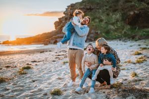 A joyful family plays and embraces on a rocky beach at sunset with ocean and cliffs in the background.