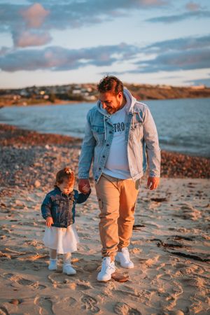 Adult and small child walking together on sandy beach at sunset with coastal waters in background.
