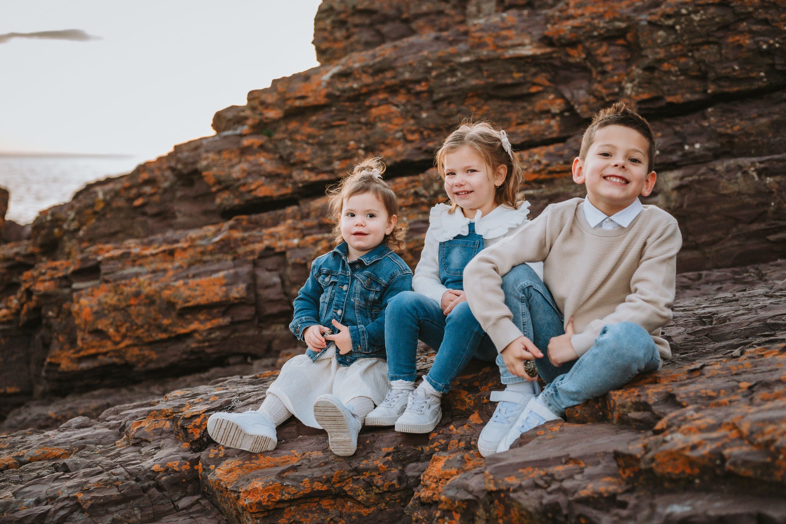 Three children sit together on reddish-brown rocky terrain wearing casual outfits in blue and beige tones.