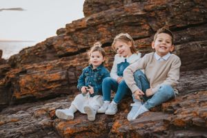 Three children in casual sweaters and jeans sit together on rocky terrain at sunset.