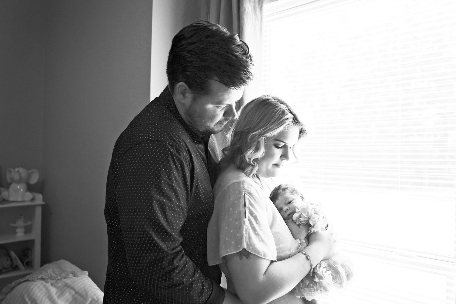 A couple stands lovingly by a window holding their newborn baby in home new session Carlsbad, Roswell, Hobbs NM
