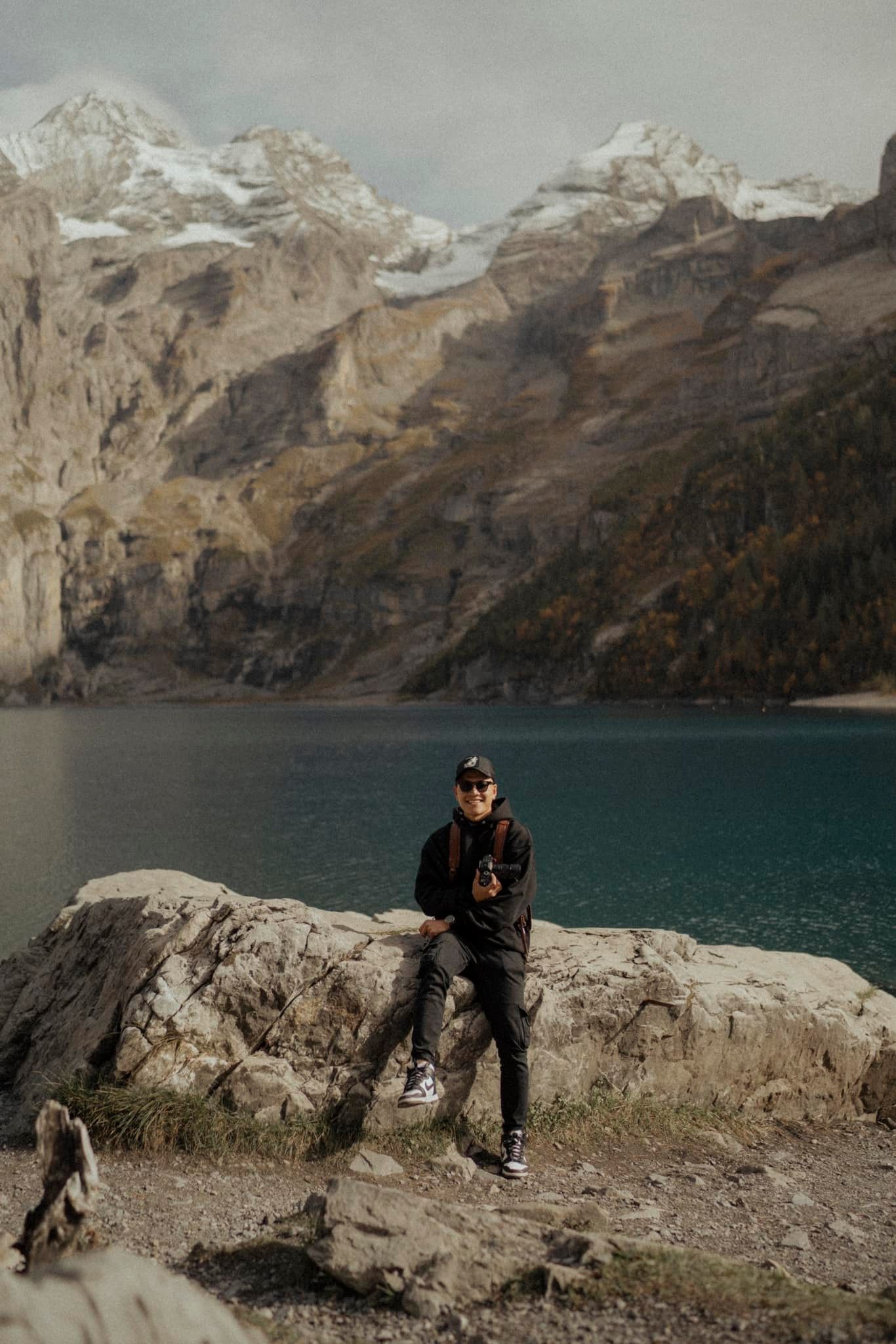 Adventure traveler poses on rocky outcrop overlooking turquoise alpine lake surrounded by mountains.