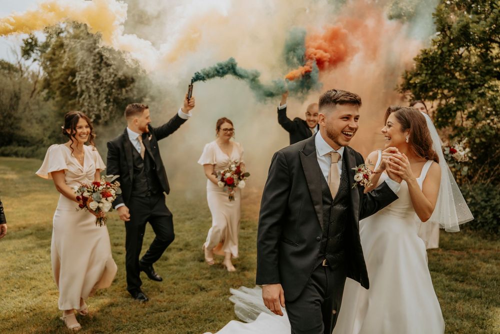 Wedding party celebrates with colorful smoke bombs in an outdoor garden setting during golden hour.