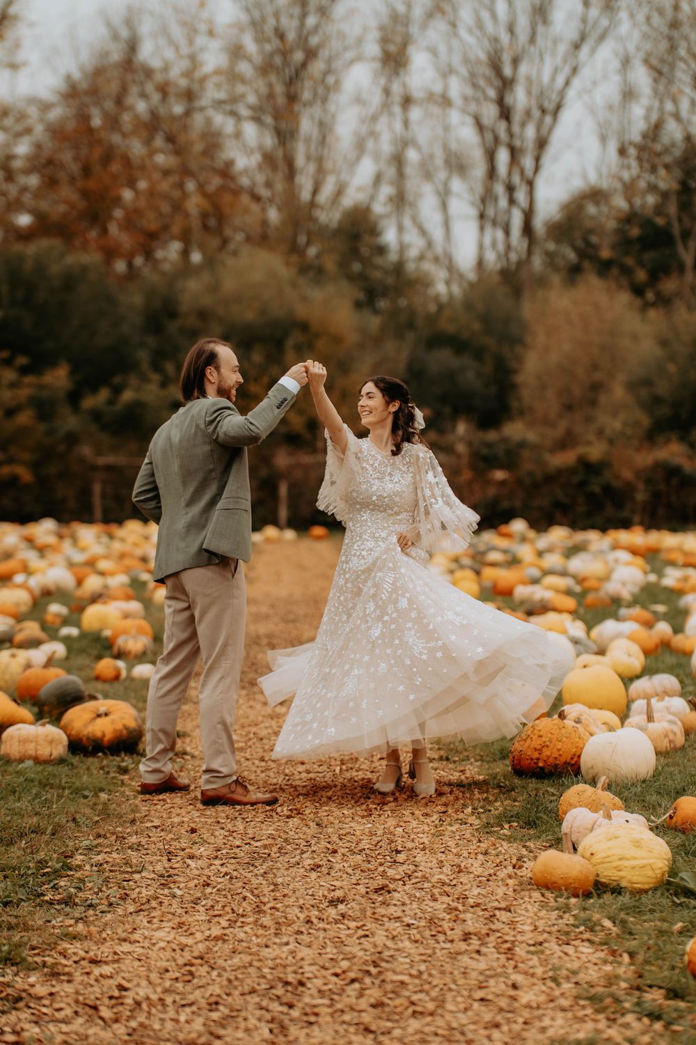 A couple dances playfully in a pumpkin patch during autumn, with the bride's white dress twirling through fallen leaves.