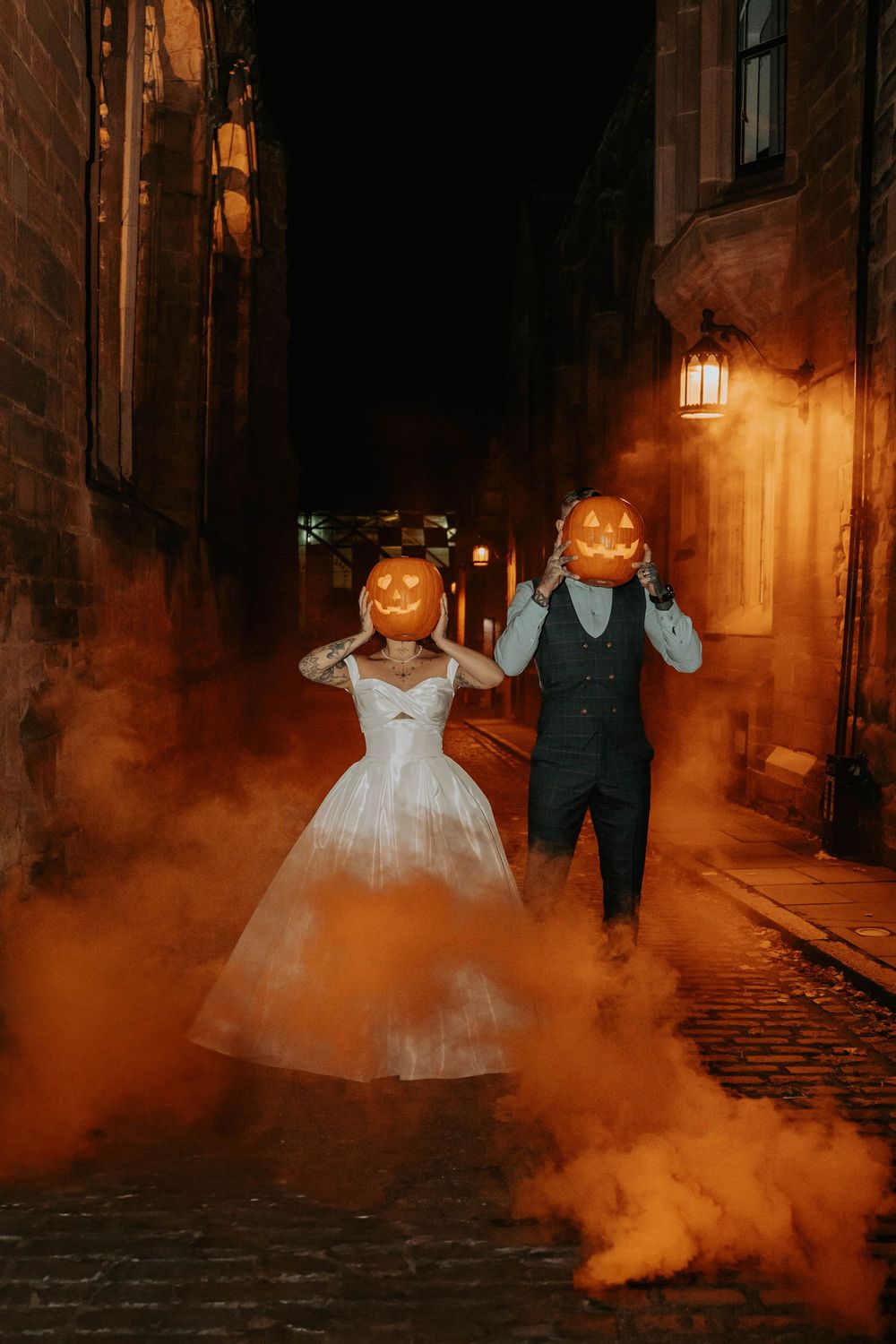 A couple in wedding attire with pumpkin heads pose on a cobblestone street with orange smoke for Halloween-themed photos.