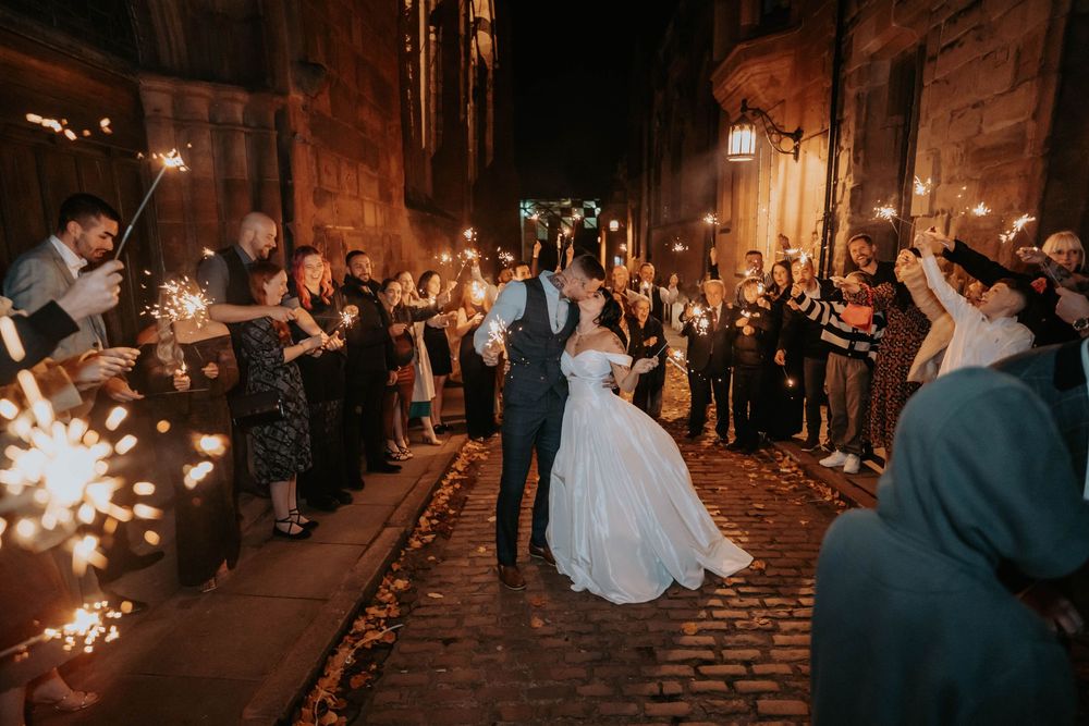 Wedding couple and guests hold sparklers during nighttime celebration on cobblestone street.