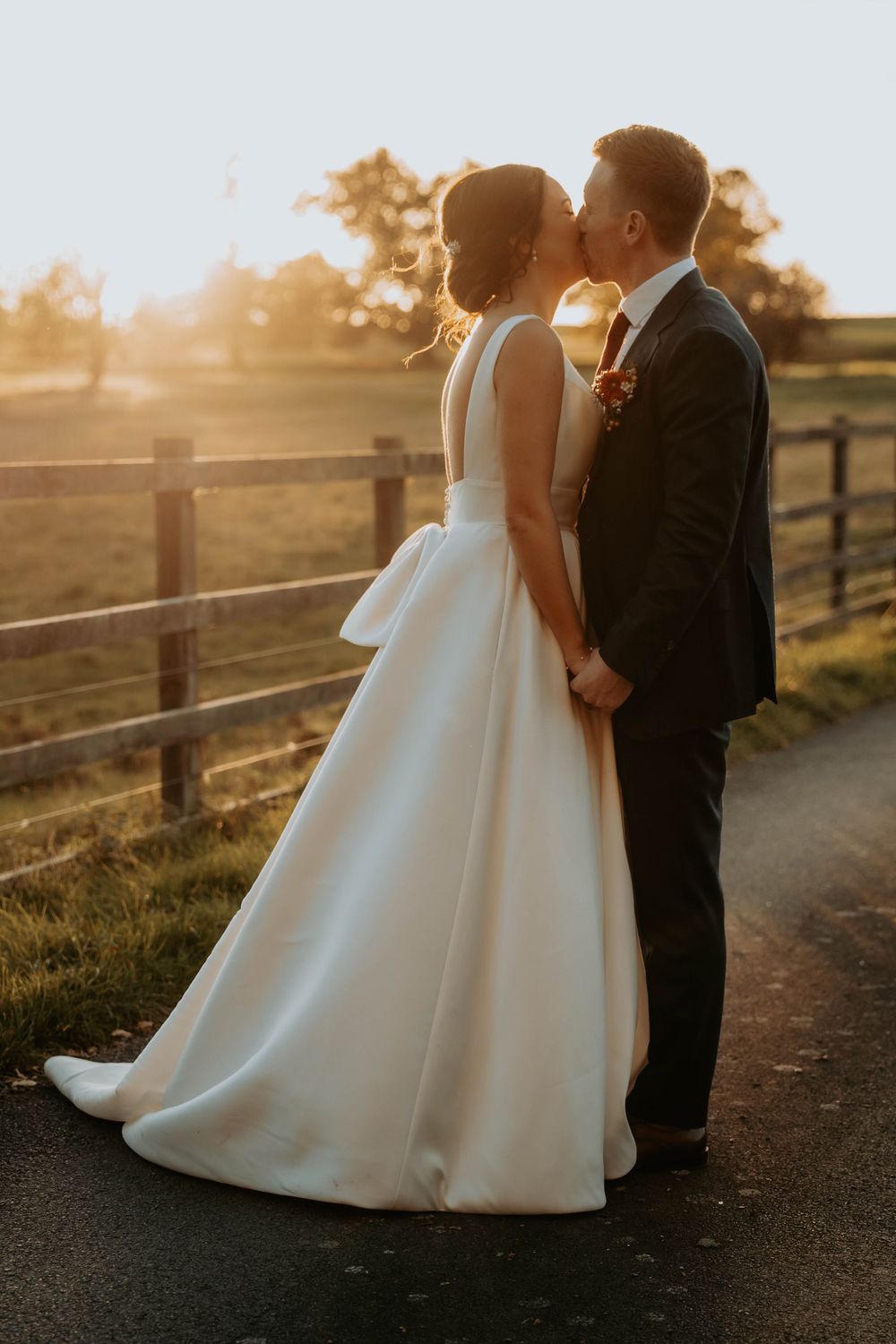 A couple in wedding attire stands together along a rustic fence at golden hour sunset.