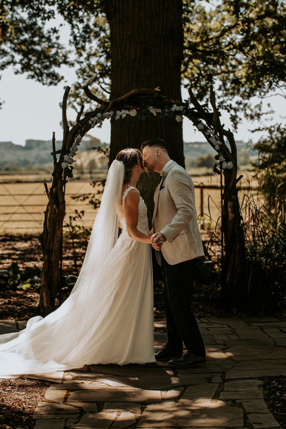 A romantic wedding couple shares intimate moments in dappled sunlight beneath tall trees at an outdoor venue.