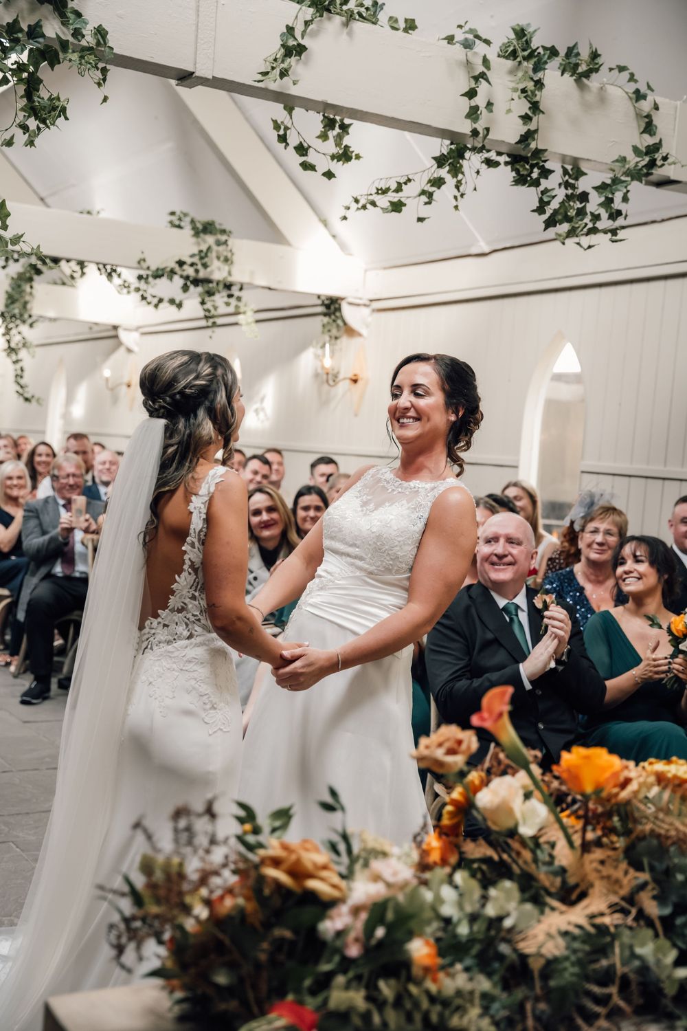 Wedding party celebrating with joyful dancing and laughter under decorative greenery at an indoor reception.
