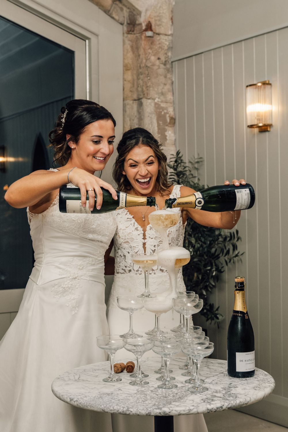 Two people in white dresses pouring champagne into tower of glasses during wedding celebration.