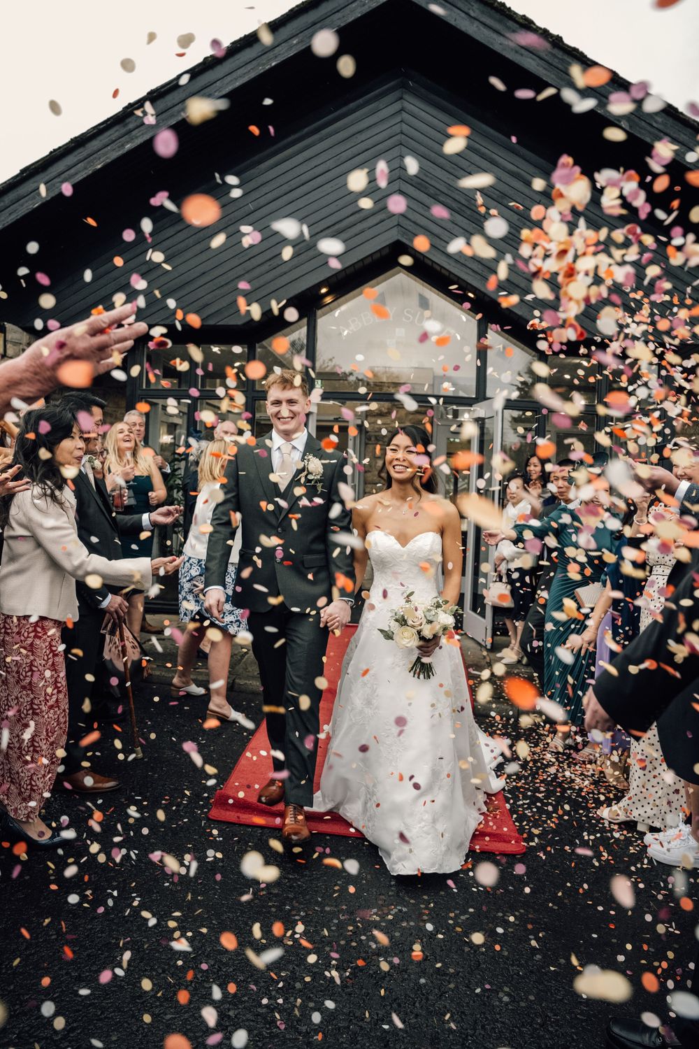 A joyful wedding celebration with confetti and flower petals being thrown outside a modern venue.