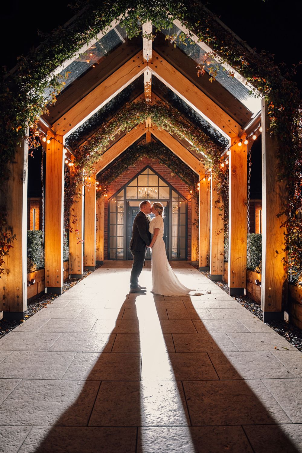 Silhouetted wedding couple in illuminated wooden archway creating dramatic shadows.
