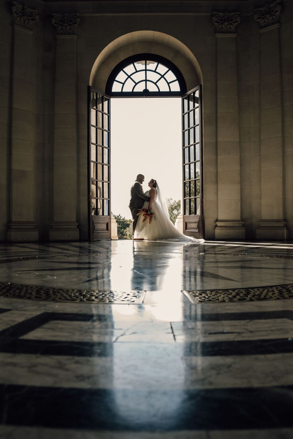 Dramatic silhouette wedding portraits in grand architectural hallway with arched windows.