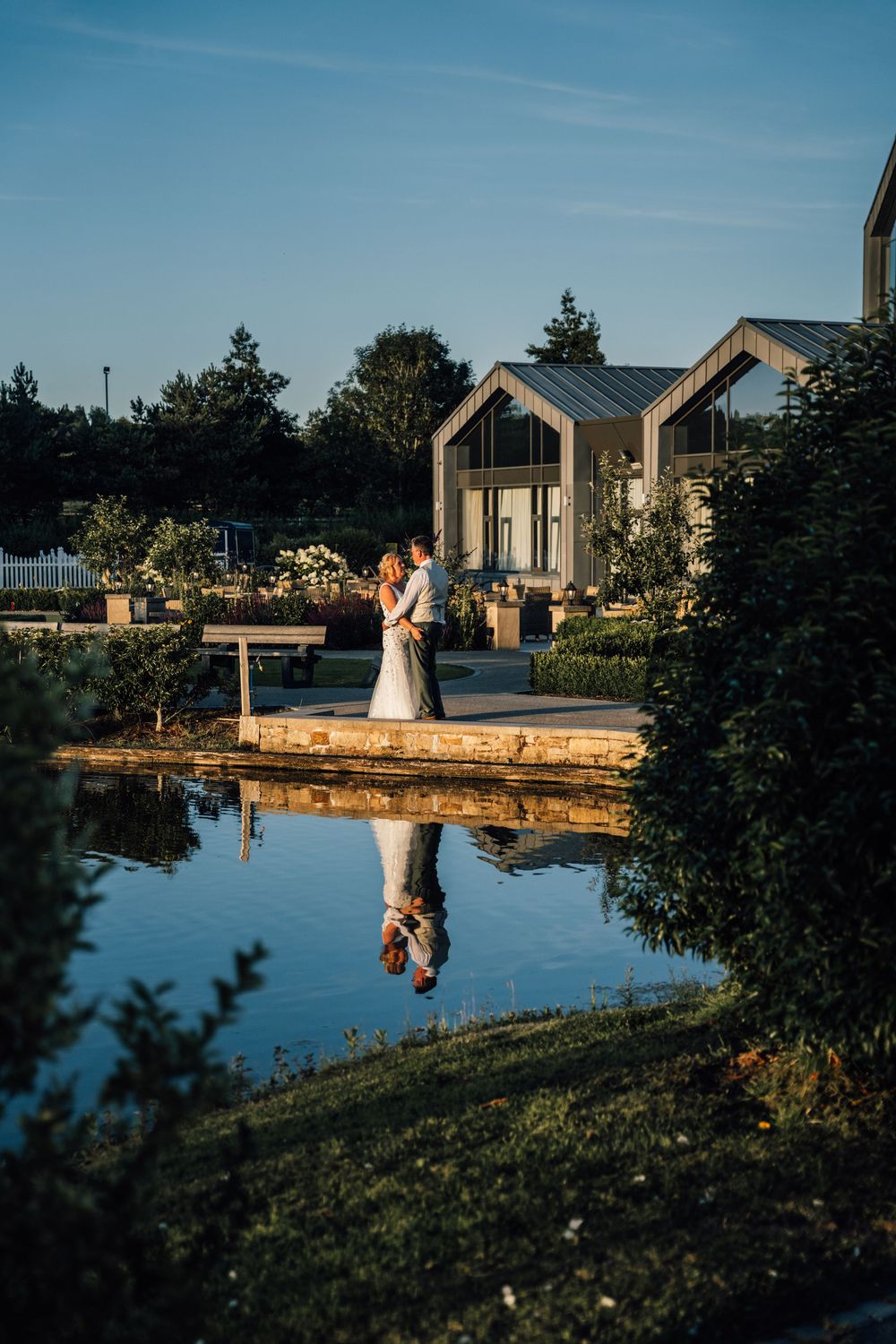 Wedding couple reflected in a serene pond with chapel architecture in the background.