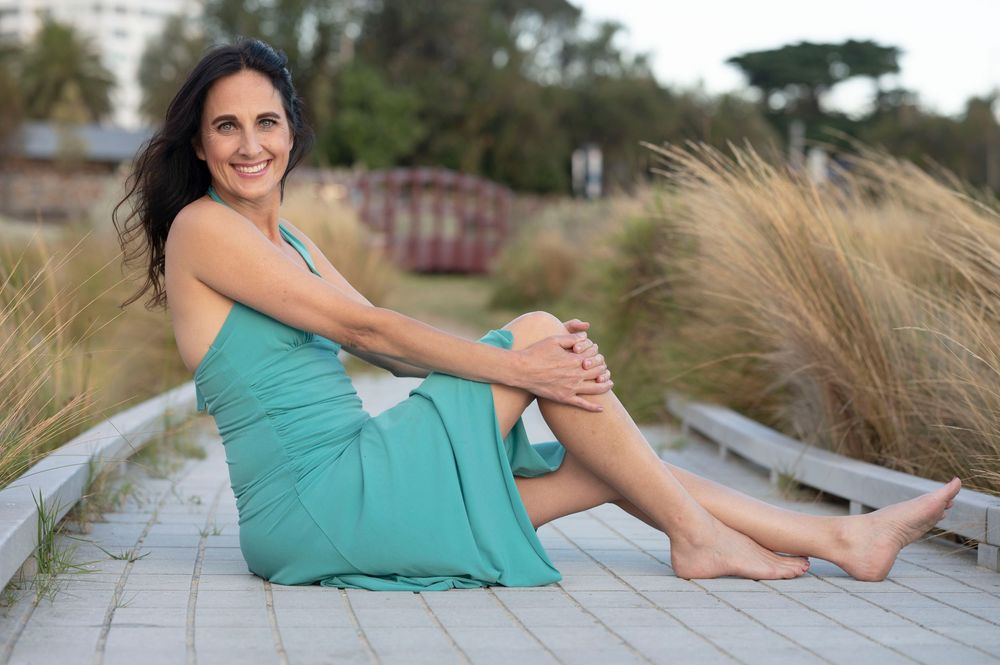 Smiling woman in teal dress sitting barefoot on a boardwalk surrounded by tall grasses.